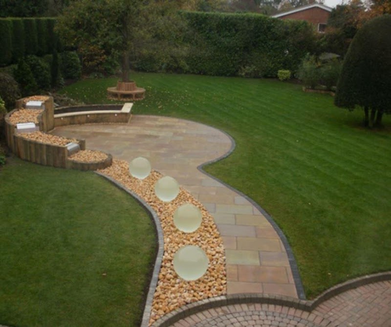 A sweeping natural-stone path and circular seating patio lit by frosted globe lights set into a cobble border, running out across a mature striped lawn.